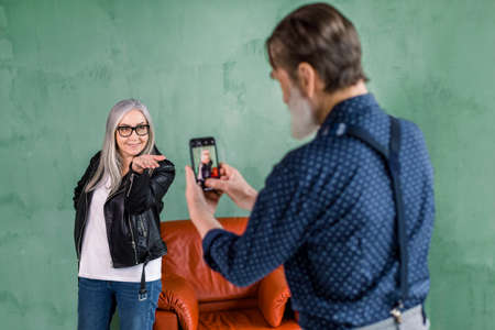 Portrait of handsome senior bearded man taking photo of his pretty woman, posing to camera on green wall background and making air kiss.の写真素材