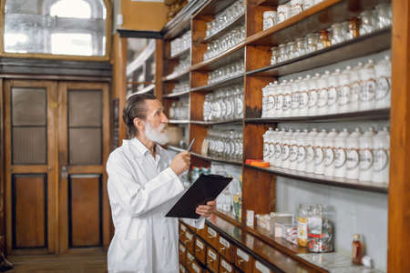 Work in pharmacy. Senior pharmacist with pen and clipboard, inspecting shelves with vintage boxes of medications, working in old ancient drugstore.の写真素材