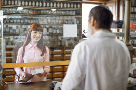 Back blurred view of middle aged man pharmacist giving medications to the young female client standing at the paydesk of the vintage old pharmacy store.の写真素材