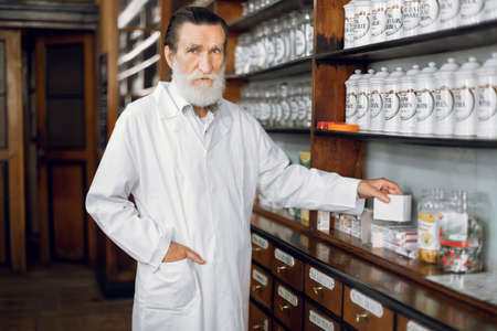 People, healthcare, pharmacy concept. Indoor portrait of pleasant professional mature bearded man pharmacist, wearing white coat, posing in beautiful old pharmacy interior.の写真素材
