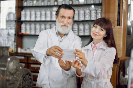 Handsome senior bearded Caucasian man pharmacist and his young female colleague, posing in vintage old apothecary, showing drug pack to camera.の写真素材