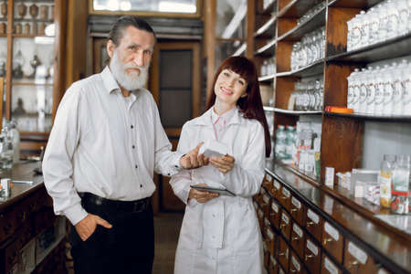 Happy senior bearded male customer standing near young Caucasian woman pharmacist, holding medicine box together, looking at camera and smiling, standing in vintage old pharmacy.の写真素材