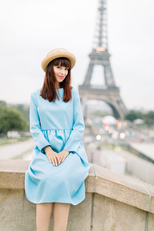 Outdoor image of gorgeous young woman in hat and blue dress sitting on stairs in beautiful European city. Paris, France, Eiffel tower on the background. Travel and people concept.の写真素材