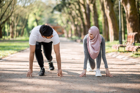 Young muslim couple in sport clothes standing in start position for run at green summer park. Man...