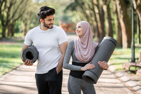 Cheerful arabian couple in sport clothes looking on each other while standing at green city park and holding yoga mats. Happy family spending free time for training outdoors.の写真素材