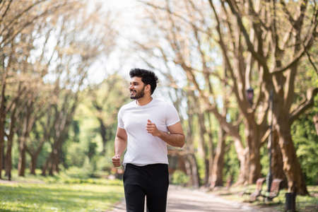 Strong muslim man in activewear running alone at green summer park. Handsome bearded guy spending leisure time for outdoors workout.の写真素材