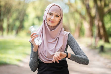 Attractive woman in sport clothes and hijab smiling and looking at camera with smart watch on hand. Young active female holding bottle of water while standing at park.の写真素材