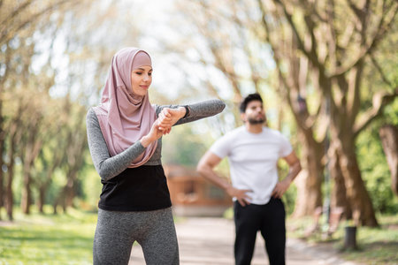 Young woman in hijab checking time on smart watch while muslim guy relaxing after training on background. Healthy sport couple exercising together at park.の写真素材