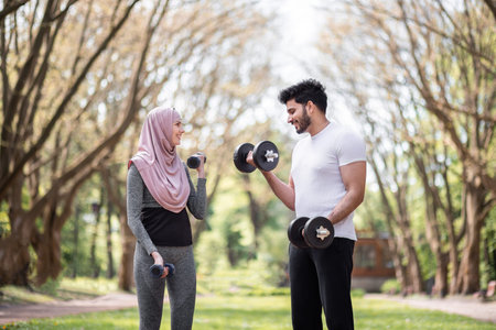 Young woman in hijab and handsome muslim man standing together at green park and training arms using dumbbells. Happy couple with sport equipment workout on fresh air.の写真素材