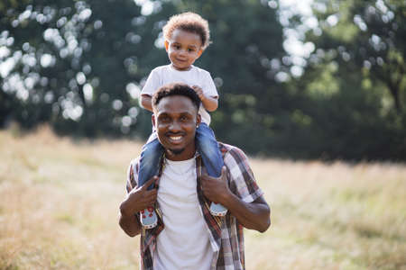 Happy little african boy sitting on fathers shoulders during walk on fresh air. Young father spending free time with lovely son on nature during summer days.の写真素材