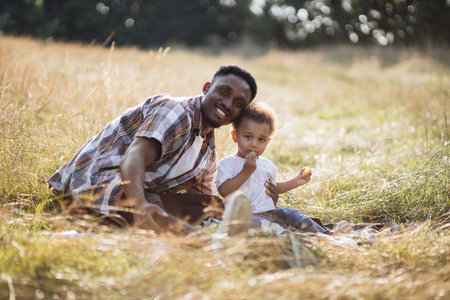 African american father embracing little son and smiling on camera while sitting together on summer field. Cute boy eating sweet grape during picnic with caring dad.の写真素材