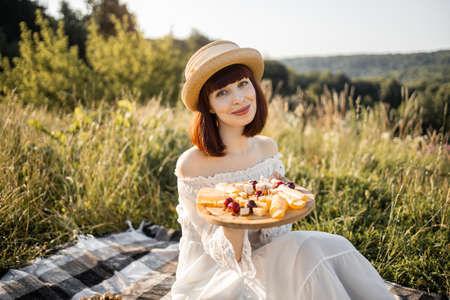 Portrait of cheerful happy young lady in dress and hat, sitting on the picnic blanket with round wooden plate with cheese and sweet cherries, enjoying vacation timeの写真素材
