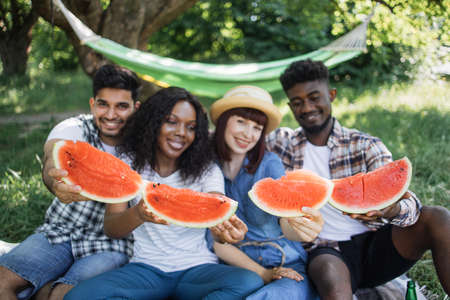 Portrait of positive young people in casual wear holding slices of sweet watermelon while relaxing together at summer garden. Concept of friendship, vacation and enjoyment.の写真素材