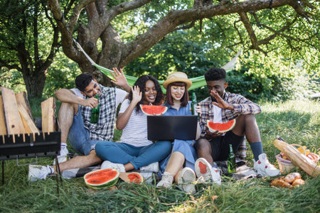 Four multiracial friends smiling and waving during video call on modern laptop while relaxing on...