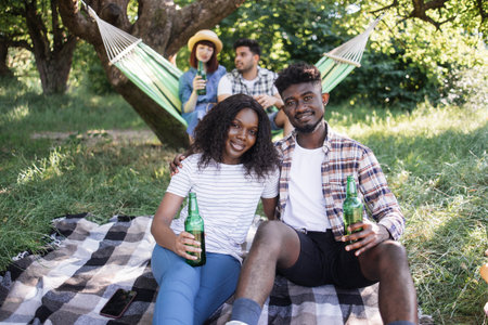 Positive black couple drinking beer and hugging while sitting on plaid outdoors. Two friends on background chatting in hammock. Young multiracial people having picnic.の写真素材
