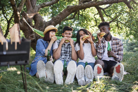 Group of four multiracial friends in summer clothes sitting at green park and eating handmade...