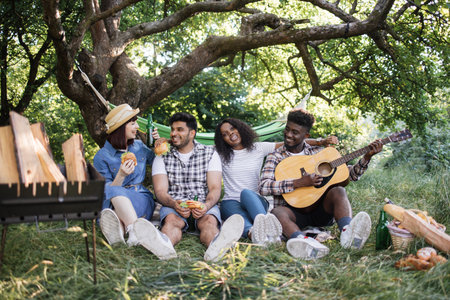 Two multiracial couple singing and playing guitar while resting outdoors. Happy friends having...