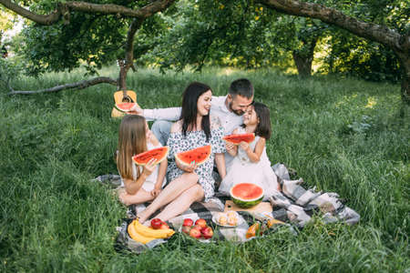 Young parents with two pretty daughters spending free time at green garden and eating tasty watermelon. Happy family having summer picnic during weekends outdoors.の写真素材