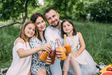 Smiling parents and their pretty daughters sitting on soft plaid outdoors and toasting with bottles...