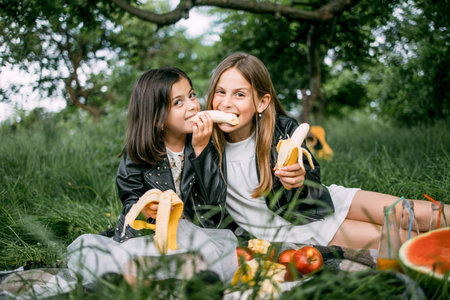 Joyful little girls in summer outfit sitting on checkered blanket and biting one banana from ends. Happy pretty sisters spending free time outdoors with fun.の写真素材