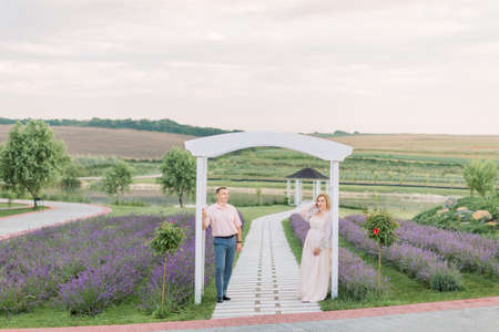 Happy together. Beautiful stylish middle aged couple in the lavender field, standing and looking away, leaning on the big white wooden archの写真素材