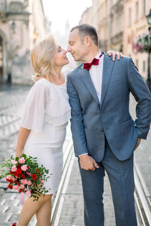 Close up portrait of loving couple, 50-aged handsome man in suit and bow tie, standing with his pretty woman in white dress with flower bouquet, on the background of old cityの写真素材