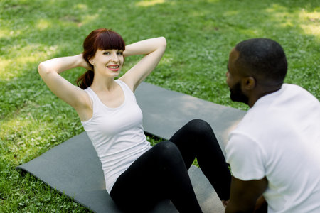 Pretty smiling young Caucasian woman exercising, doing sit-ups on yoga mat outside in green summer...
