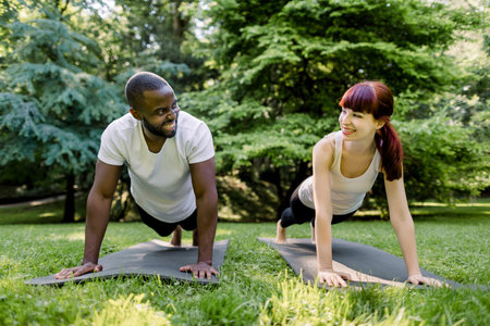 Multiethnic smiling couple, African man and Caucasian girl, doing core workout together in park. Fit...