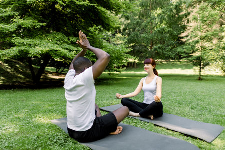 Morning yoga exercises and healthy lifestyle. Multiethnic couple doing yoga in park together, sitting in yoga pose on yoga mats outdoor at sunny summer day in park. Training and meditation.の写真素材