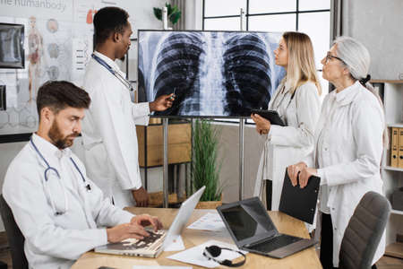 Team of medical specialists listening african american male colleague pointed on computed tomography result in clinic. Multicultural competent doctors with help of gadgets examining patient analysis.の写真素材