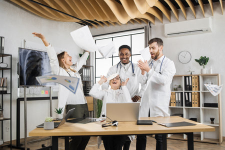 Excited multiracial scientists in white medical uniform laughing and throwing documents in air during laboratory meeting. Success teamwork and healthcare achievement concept.の写真素材