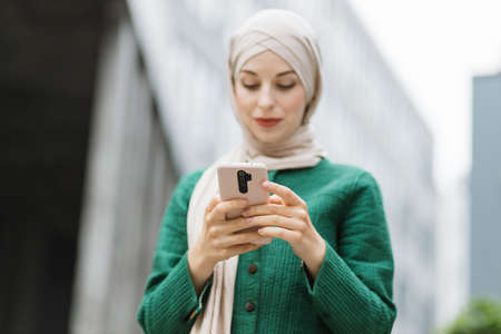 Close up of pleasant business woman in hijab using modern smartphone while standing outdoors, focus on phone. Young arabian lady typing messages on mobile outside office.の写真素材