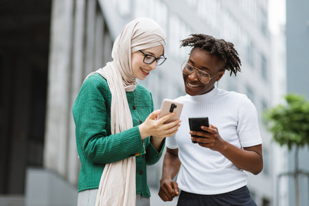 Muslim businesswoman in green jacket showing something on smartphone to her female colleague in casual clothes. Pretty african american woman standing near and holding smart phone in hands.の写真素材