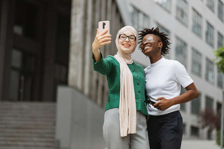 Smiling arabian woman standing on city street and taking selfie on smartphone with her african female colleague. Charming women, one in hijab and other her african american friend taking selfie.の写真素材