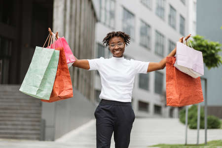 Shopping, sale, purchase concept. Young excited fashionable African woman, holding colorful shopping paper bags, posing in street with arms raised. Blurred modern city on the background.の写真素材