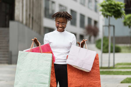 Summer shopping concept. Pretty happy smiling fashionable african woman wearing white t-shirt and dark pants posing with colorful paper bags outdoors in the city street. Copy space.の写真素材