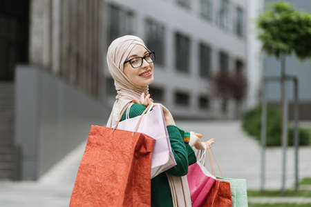 Close up portrait of beautiful young muslim woman, wearing casual clothes, going for shopping in background of modern city buildings, posing to camera with shopping bags. Copy space.の写真素材