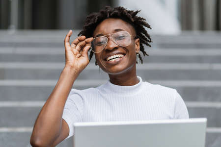 Charming business woman in stylish elegant clothes sitting on stairs outdoors and using wireless laptop. African American female working on distance.の写真素材