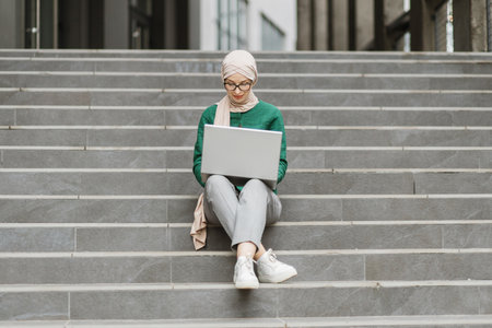 Charming woman in stylish elegant clothes and hijab sitting on stairs outdoors and using wireless laptop. Muslim female working on distance.の写真素材