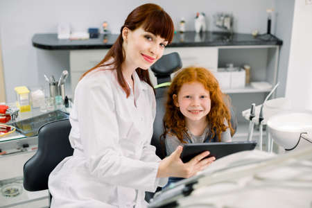 Medicine, pediatric dentistry and oral care concept. Female smiling dentist showing tablet pc computer to happy kid patient, curly red haired school girl at modern dental clinic.の写真素材