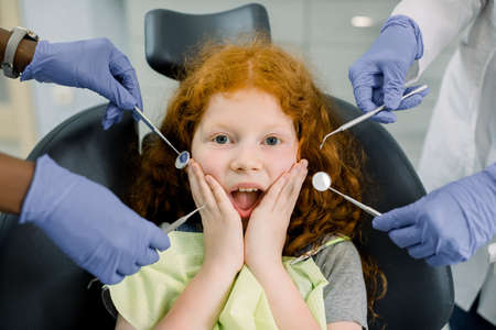 Healthy teeth, caries prevention and pediatric dentistry. Close up of scared funny little red haired curly girl, looking at camera and screaming and hands of two dentists in gloves with dental tools.の写真素材