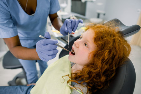 Side close up view of curly red haired girl lying on dentist chair with open mouth while female...