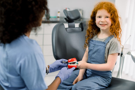Happy smiling kid at dentists office. Little girl with red curly hair, sits in dental chair and...