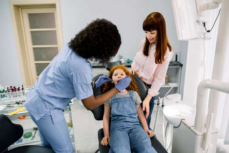 Young female African American dentist with dental tools, making dental check up of her little patient girl. Young mother reassuring her frightened cute daughter during examination.の写真素材