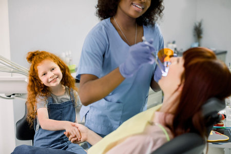 Young little red haired curly girl supporting her mom at dentists office during checkup. Female...