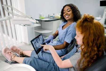 Little patient kid girl with curly red hair sitting in dentistry chair and pointing on the tablet with teeth panoramic radiography. Pretty african woman doctor explaining the ways of treatment.の写真素材