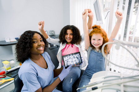 Smiling happy children, two multiracial girls, visiting dentist, sitting in dental chair, gesturing...
