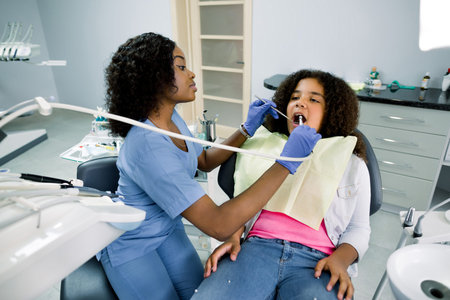 Shot of young pretty female african dentist in blue uniform, making dental check up and caries treatment with drill for her cute little patient, mixed raced curly girl, sitting in dental chair.の写真素材