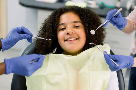 Charming little African American girl afro hair, sitting in dental chair, smiling and looking at...