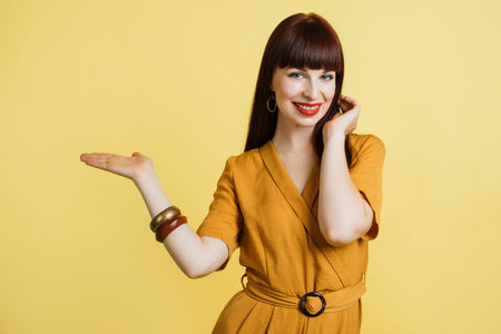 Portrait of beautiful girl in yellow dress, showing imaginary something, empty copy space for some text, product or slogan, on the open hand palm, isolated on yellow background.の写真素材